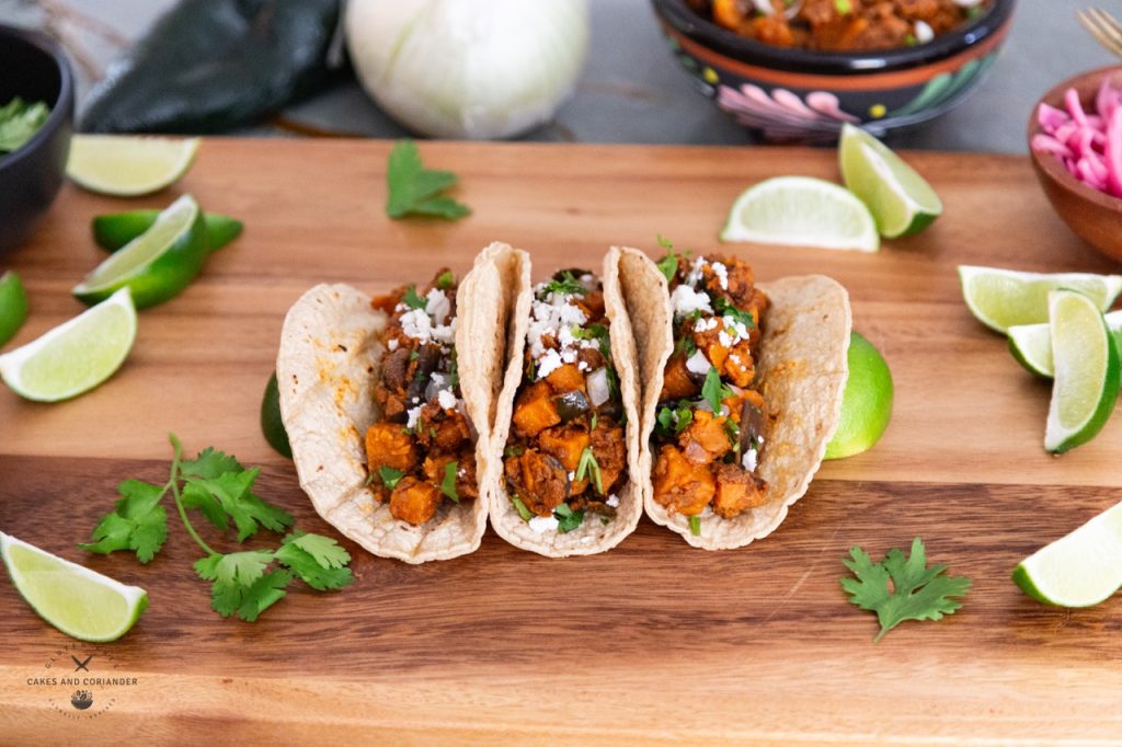 Cutting board with three sweet potato poblano tacos with limes, cilantro, queso fresco, pickled red onions.