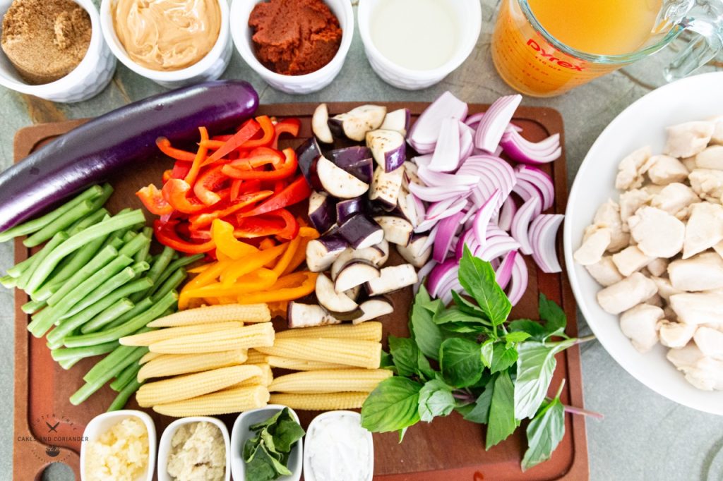 A variety of colorful vegetables chopped and ready for cooking sitting on a wooden cutting board. Vegetables include red bell peppers, baby corn, thai basil, onions, eggplant, and babmoo shoots